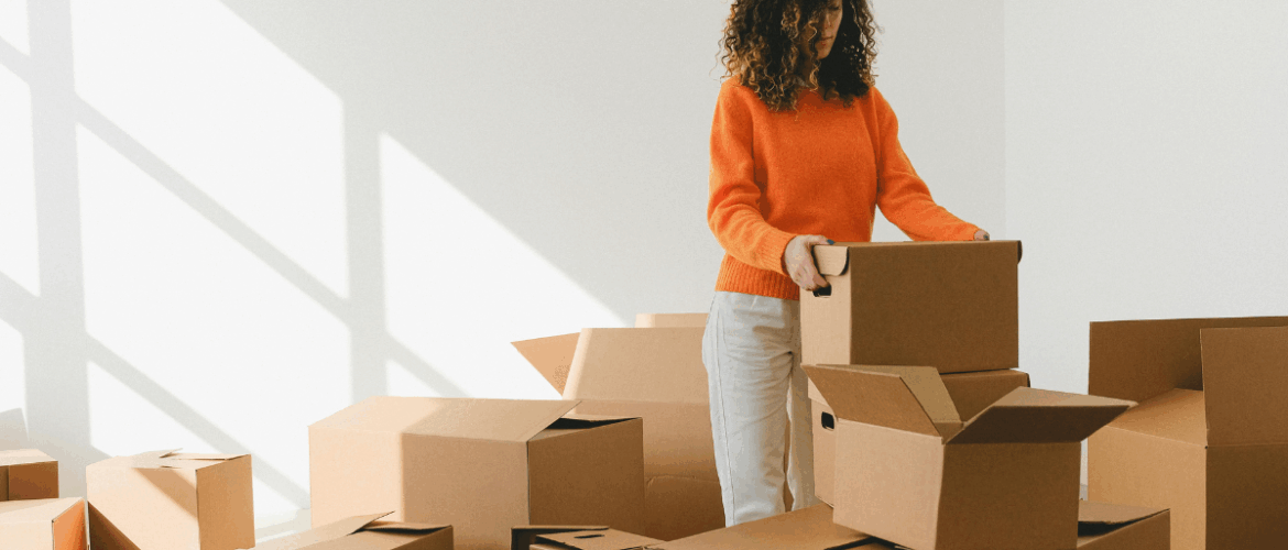 uma mulher negra de suéter laranja está em pé organizando caixas de papelão, simbolizando a mudança para desocupação ou ocupação de um imóvel